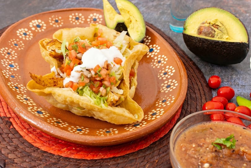 Chicken taco salad in a fried tortilla bowl with salsa, avocado, and cherry tomatoes.