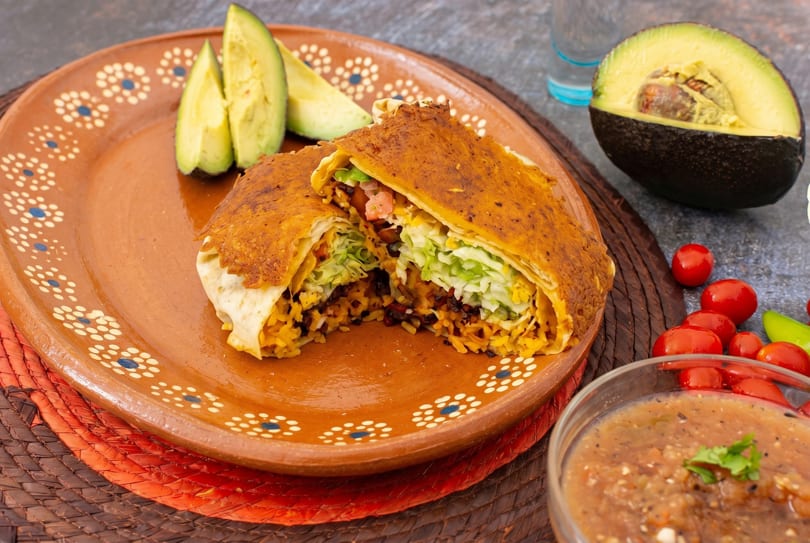 A crispy burrito, cut in half, revealing rice, beans, and lettuce, served with avocado slices and salsa.